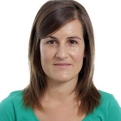 Alba is pictured smiling in front of a white background. She has long brown hair and is wearing a teal top.