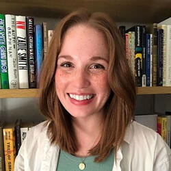 Allie is a white woman with medium-length reddish hair, wearing a green tank top and white button-up shirt. She is standing in front of a full bookshelf, looking directly into the camera and smiling.