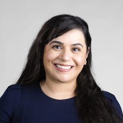 Ayesha is pictured smiling in a professional photograph in front of a gray background. Ayesha has long brownish black hair and is wearing a blue top.