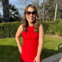 Beatriz is pictured standing and smiling outdoors. In the background is a patch of grass, hedges, and trees. Beatriz has long brown hair and  is wearing sunglasses and a red dress.