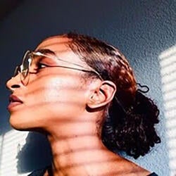 Brianna is pictured in a facial profile as she looks to the left of the photograph. In the background is a textured wall with some light patterns from a window. Brianna has long brownish black hair and is wearing glasses with a metal frame.