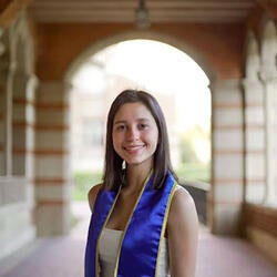 Emmi is pictured here under an arched pathway, smiling and wearing some graduation regalia. She has long brown hair, is wearing a white top.