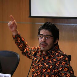 Fernando is pictured here presenting a talk about decolonialiy,  raising his right hand to emphasize his speech. He is wearing a long-sleeve orange and green shirt. He has black curvy hair and he wears glasses. He is sitting down.