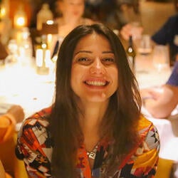 Jovana is pictured smiling at a formal event. The background is blurred, and in the foreground, Jovana is in focus.. She has long brown hair and  is wearing a silver-toned necklace and a colorful top with splashes of orange, yellow, black, and white.