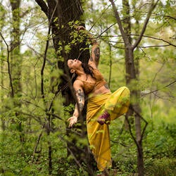 Marielys, an Afro-indigenous brown skin woman dancing between trees in the land of the Munsee and Canarsee tribes of the Lenape peoples. She wears a lace sleeveless crop, loose pants, and sneakers. Balancing one leg, the other suspended in the air in front of her bent at the knee. Her torso is slightly bent sideways, long hair cascading, one hand reaching the Earth palm facing up, the other hand above her head palm down. Marielys looks at the open sky, dreaming with the intangible. Photo by Rachel Keane.