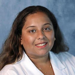 Marina is pictured here in a professional photograph in front of a gray background. She is smiling and wearing a white lab coat with the UC Riverside School of Medicine logo.. She has long brown hair and is wearing a gold-toned necklace.