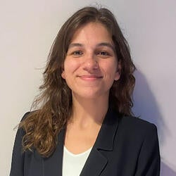 Martina is pictured smiling in front of a white background. She has long curly brown hair and  is wearing a black blazer over a white top.