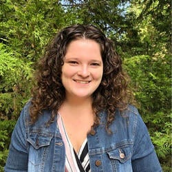 Mikaela is pictured standing and smiling outdoors, with trees in the background. She has long curly brown hair, a bright smile, and is wearing a jean jacket and a pink, white and black striped shirt.