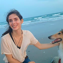 Paula is pictured here at the beach, smiling while kneeling with a hand on her dog Zoe, a collie. Paula has long brown wavy hair, is wearing a tan tee shirt and jeans.