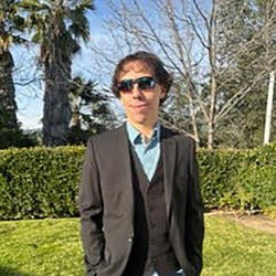 Robert is pictured standing and smiling outdoors. In the background is a patch of grass, hedges, and trees. He has medium-length curly  brown hair and is wearing a dark gray three-piece suit with a blue collared shirt underneath.