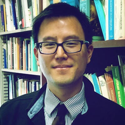 Asian American man with glasses in a collared shirt, tie, and blazer in front of a bookcase
