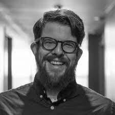 Black-and-white photo of a white man with glasses and a beard smiling in a hallway
