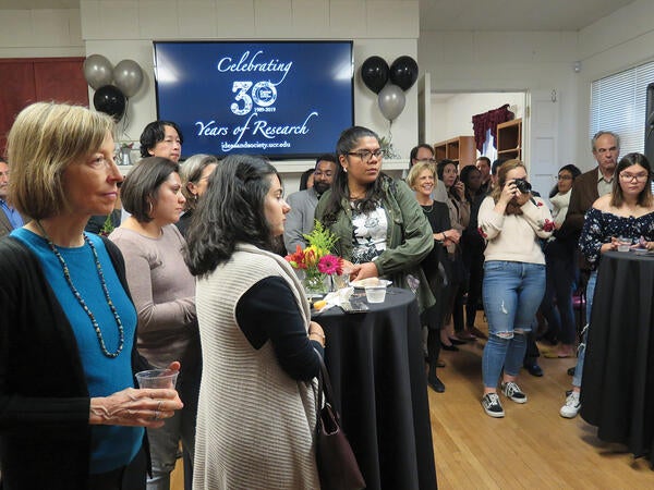 A diverse group of people standing around tables with a screen that says "Celebrating 30 Years of Research"