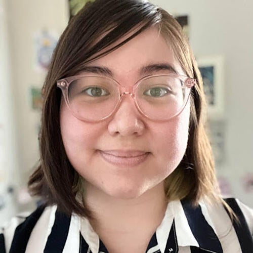 Loren has short, shoulder-length brown hair and clear frame glasses. They are wearing a black and white striped button-up shirt. They are standing in front of a white wall, looking into the camera and smiling.